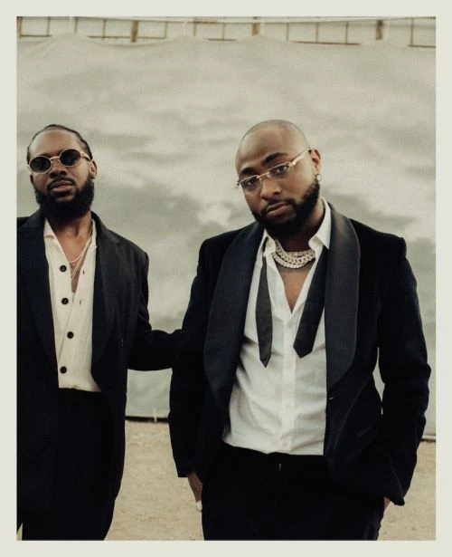 Two adult males pose in formal black suits against a textured cloudy background. One man wears round glasses; the other a chain necklace.