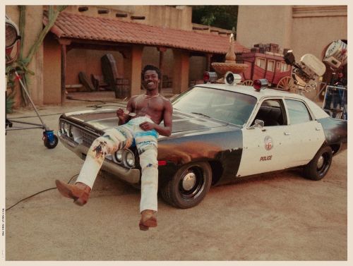 Shirtless man with sunglasses sits on a vintage police car outside a desert building with film equipment visible, under bright light.