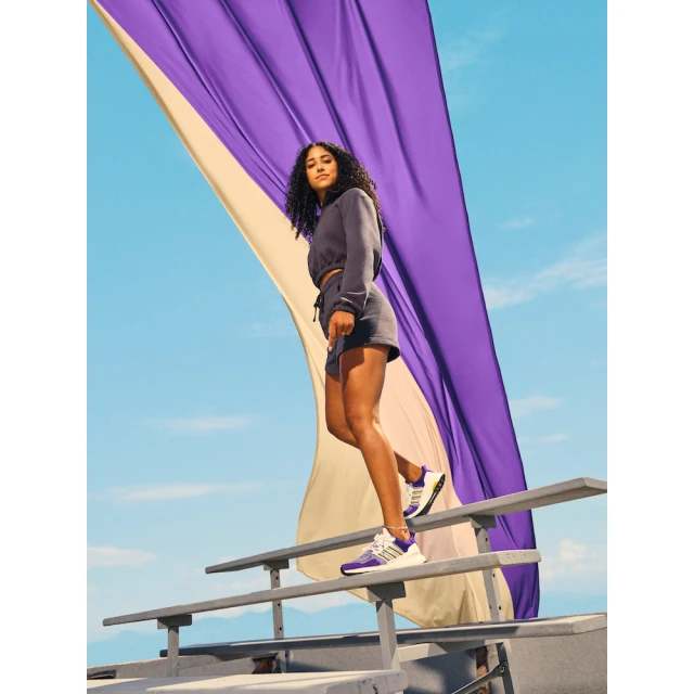 Woman in athletic wear poses on steps against a sky backdrop with draped fabrics, showcasing lifestyle fashion and athletic footwear.
