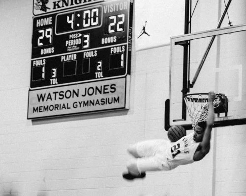 Basketball player hanging from net after dunking with game scoreboard showing Watson Jones Memorial Gymnasium in the background.