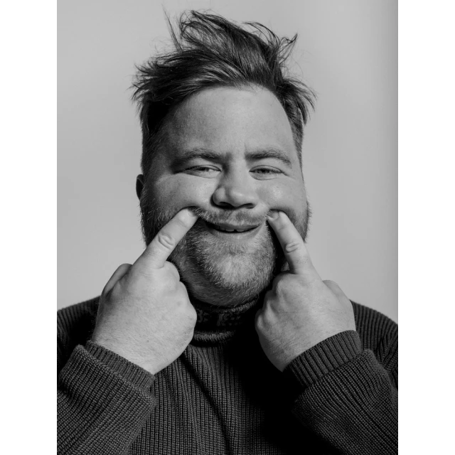 Black and white portrait of a cheerful adult male with disheveled hair, smiling broadly while using his fingers to pull up the corners of his mouth.