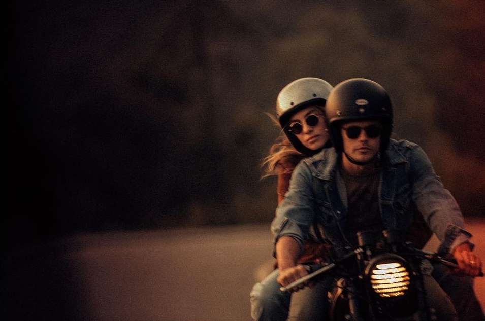 A man and woman ride a motorcycle on a road. The couple wears helmets and casual clothing, with a blurred background of trees and sky.