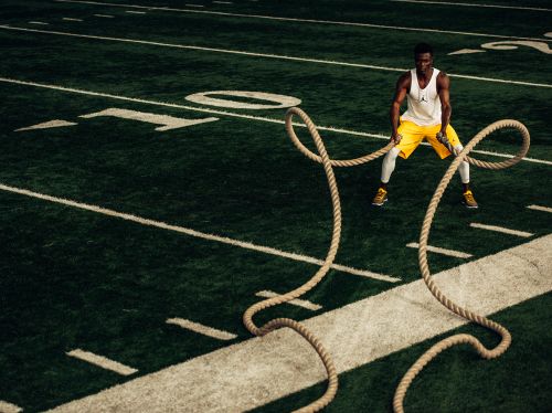 Athletic African American man in activewear doing workout using battle ropes on green football field with white lines. Outdoor fitness.