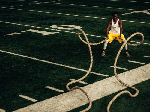 Athletic African American man in activewear doing workout using battle ropes on green football field with white lines. Outdoor fitness.