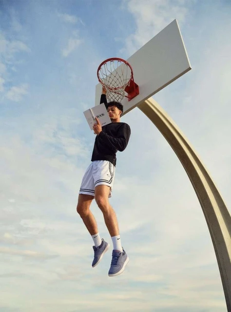 Young man reading a book on a basketball hoop, wearing athletic shorts, socks and sneakers, against a blue sky background.