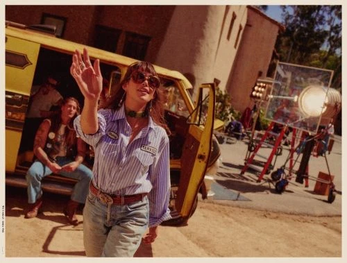 Woman in striped shirt waves near a yellow van with film crew, camera lights, and equipment in an outdoor production setting.