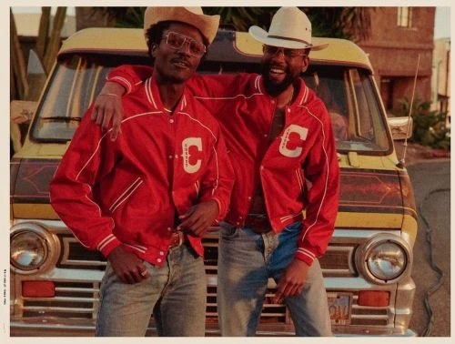Two adult men in vintage-style red jackets and cowboy hats pose in front of a yellow van. Retro, warm-toned outdoor setting.