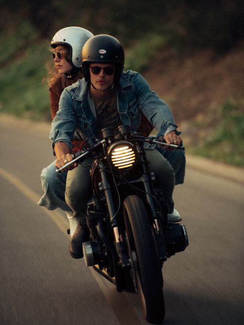 Couple riding a vintage motorcycle on a scenic road, wearing helmets and casual attire in the evening light, trees visible in background.