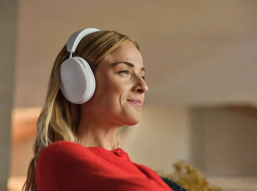 A woman with light blonde hair and a red sweater is listening to music with white headphones indoors with a blurred background.