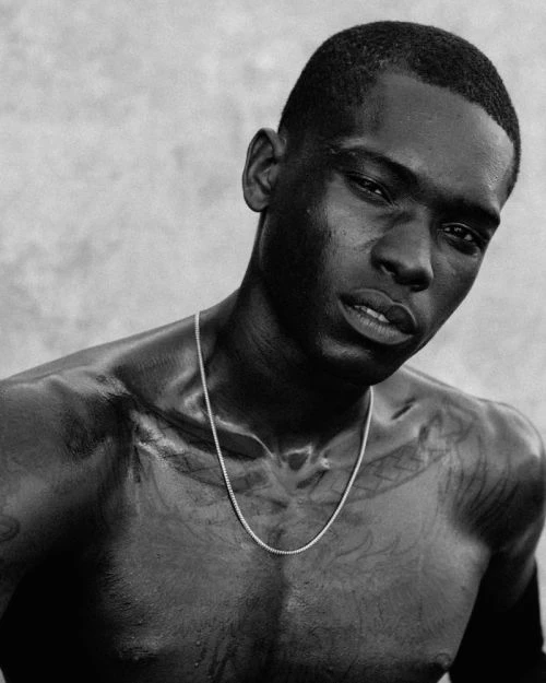 Striking black and white portrait of an African American male model with a shaved head, wearing a silver chain against a textured wall.