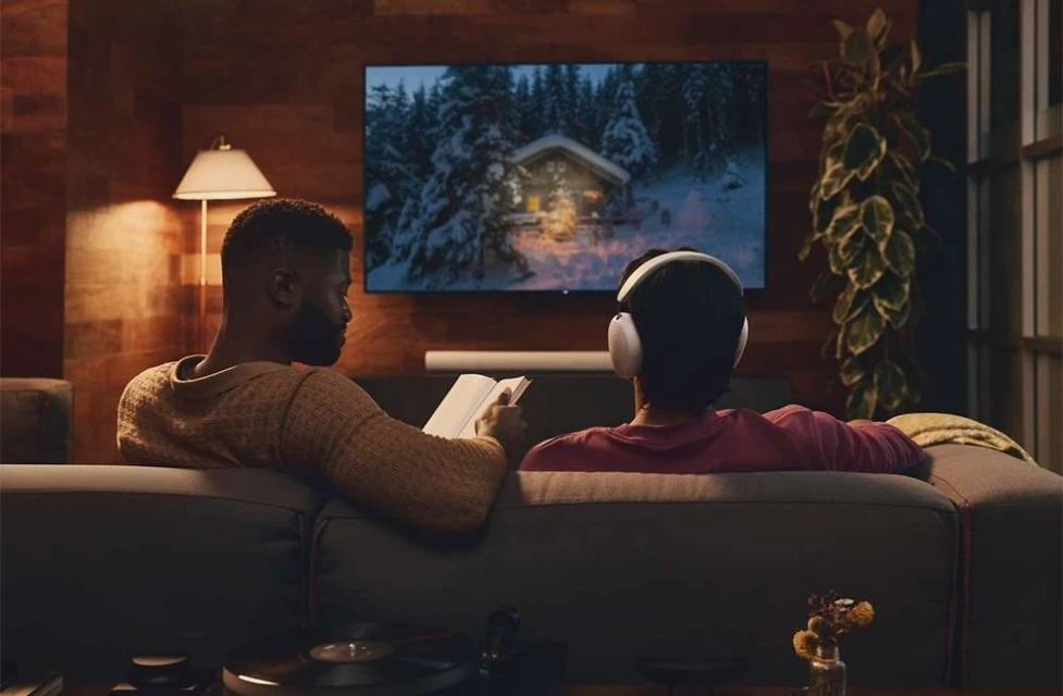 Two men relaxing on a couch, one reading a book and the other listening to headphones, with a TV displaying a winter cabin scene.