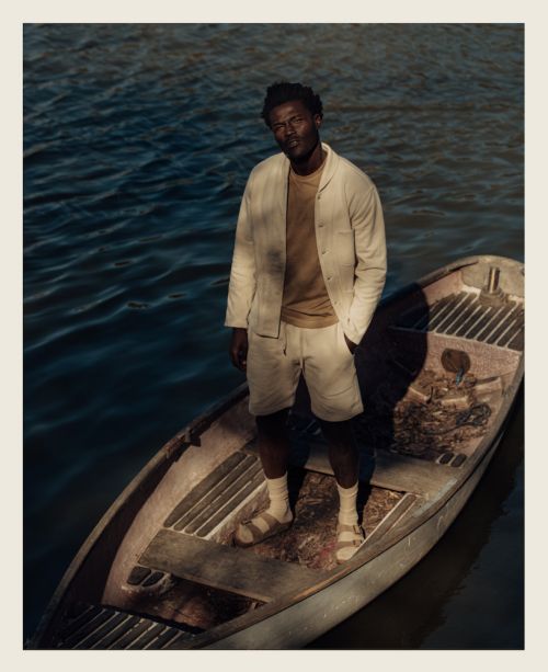 African-American man poses in a boat on the water wearing a jacket, shorts, t-shirt, socks, and sandals on a sunny day.