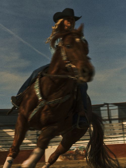 A blonde woman in a cowboy hat rides a brown horse in a rodeo arena, horse's tail blowing behind, dusty environment, action shot.