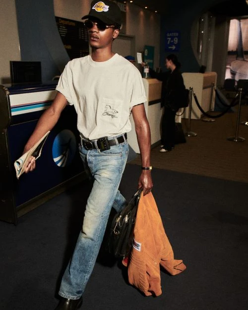A young adult male walks through an airport in casual clothes, holding a newspaper and jacket, with gate signage in the background.
