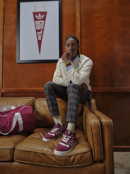 Young adult sits on brown leather couch indoors with maroon Adidas shoes and matching duffel bag. Framed collegiate pennant on wall.
