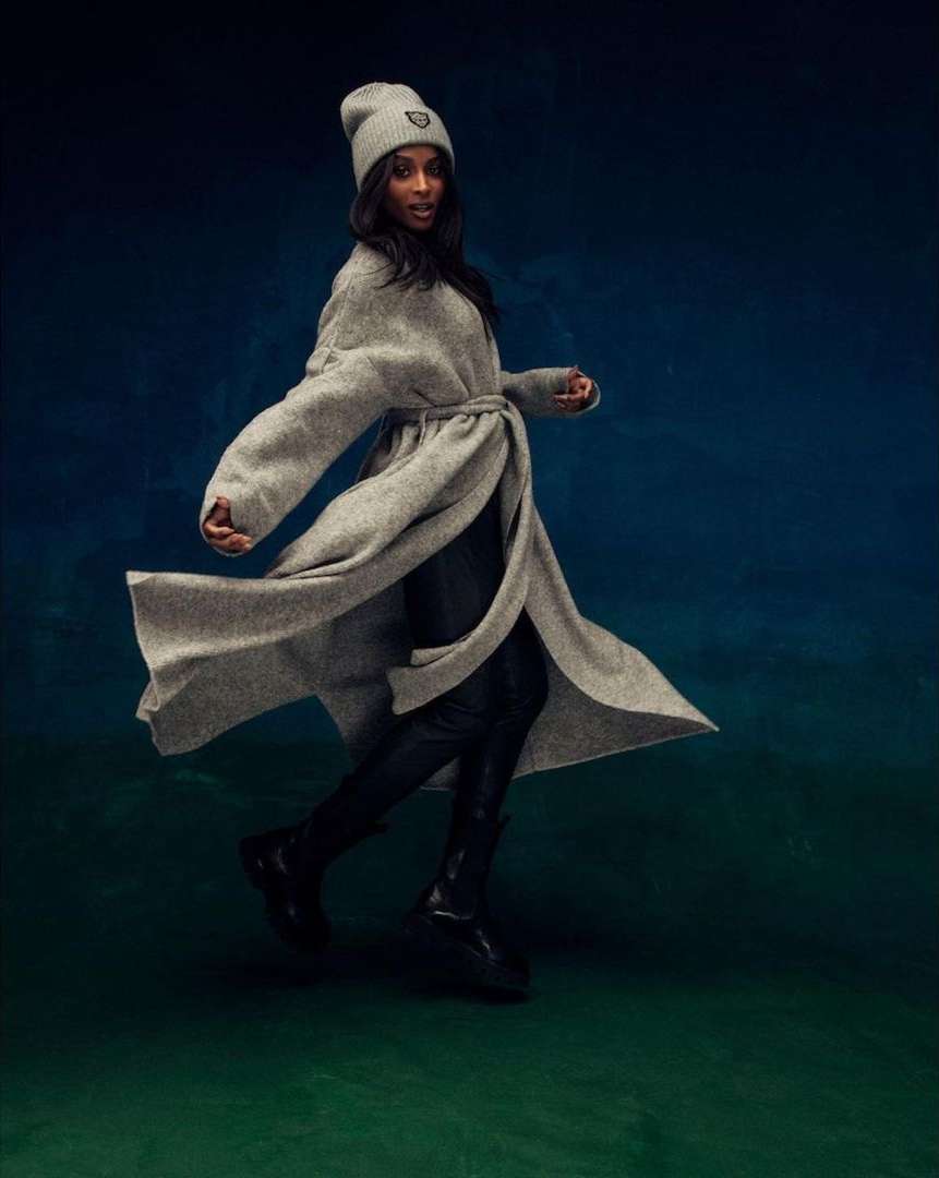 Fashionable woman in motion, wearing a grey wrap coat and beanie with black boots, against a blended blue and green backdrop.