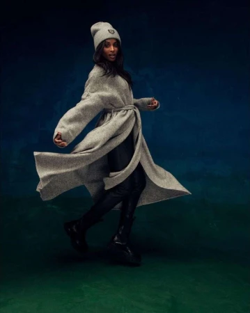 Fashionable woman in motion, wearing a grey wrap coat and beanie with black boots, against a blended blue and green backdrop.