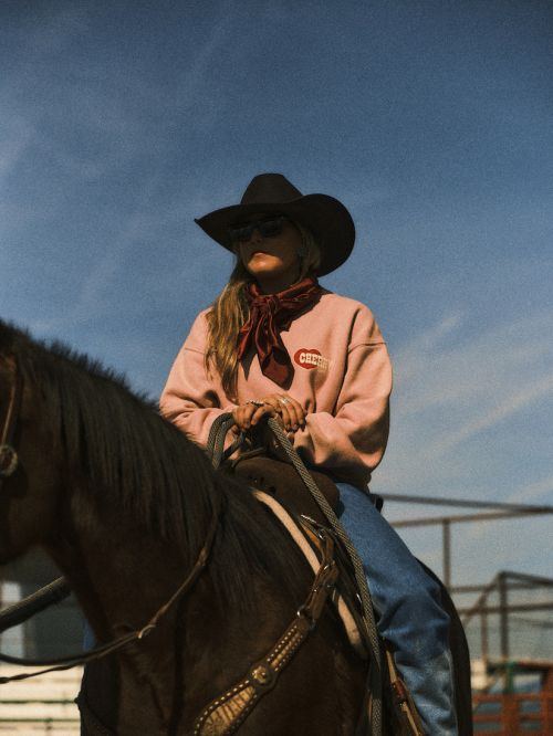 Young adult woman riding a dark horse outdoors wearing cowboy hat, jeans, and a pink sweatshirt with a patterned scarf. 