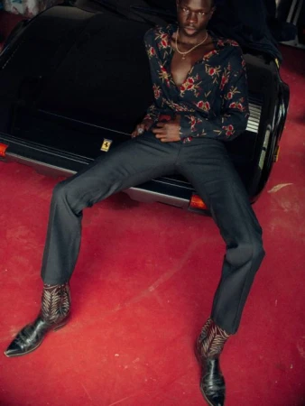 Man wearing a floral patterned shirt and dark trousers leaning against a black car over a red carpet background