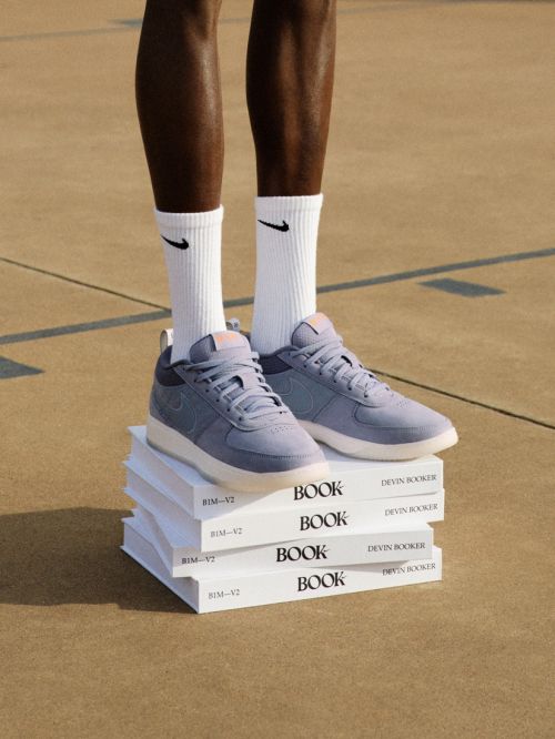 Nike shoes are stacked on a pile of books next to dark-skinned human legs wearing Nike socks, all standing on a concrete surface.