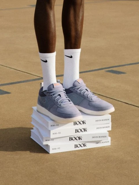 Nike shoes are stacked on a pile of books next to dark-skinned human legs wearing Nike socks, all standing on a concrete surface.