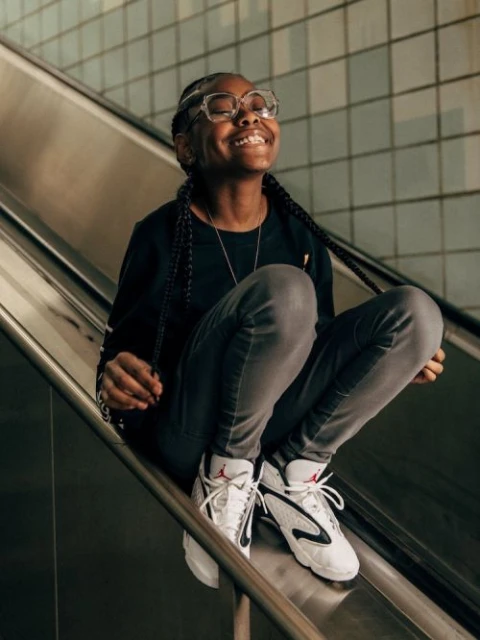 Smiling young African-American woman wearing glasses and sneakers, sitting on a silver metal escalator in an urban environment.