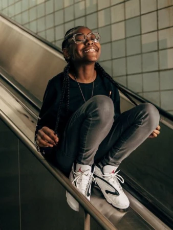 Smiling young African-American woman wearing glasses and sneakers, sitting on a silver metal escalator in an urban environment.
