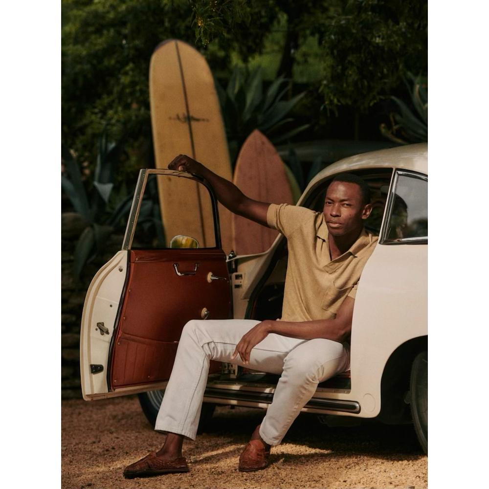 A man sits in a vintage car with the door open, wearing a tan polo shirt, light-colored pants, and loafers; surfboards in the background.