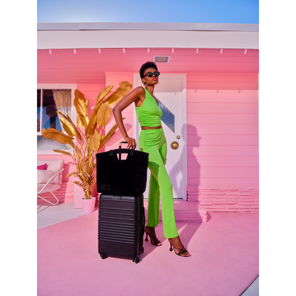 Woman poses in front of a pink house with luggage and a tote bag, wearing sunglasses and a lime green two-piece outfit, sunny day.