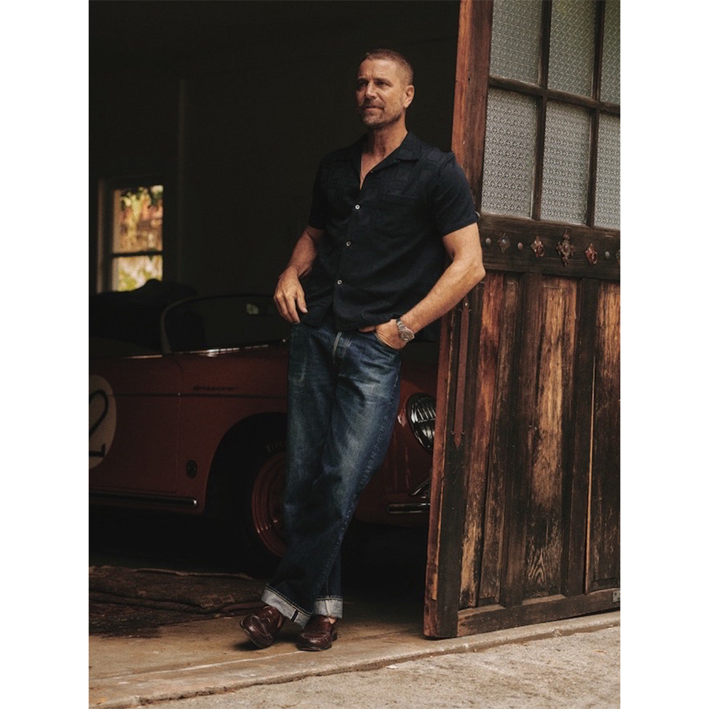 Adult man poses against a barn door next to a red sports car, wearing a black button-down shirt and jeans, showcasing casual style.