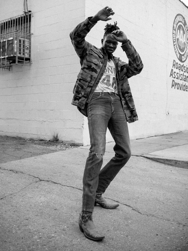 A young adult stands on a city sidewalk wearing a camouflage jacket, jeans, and cowboy boots, with a white building in the background.