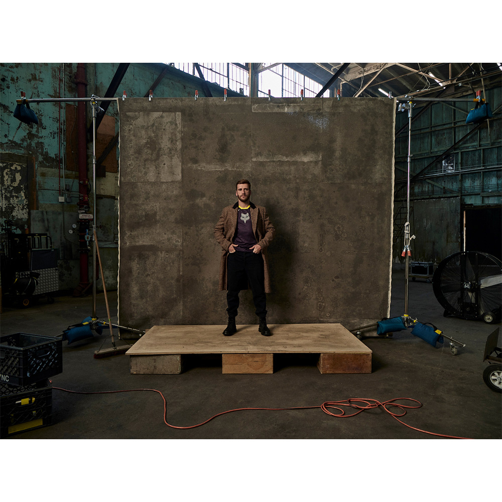 Full-length shot of an adult man standing on a wooden platform, wearing a jacket, t-shirt, and pants against a textured grey wall backdrop.