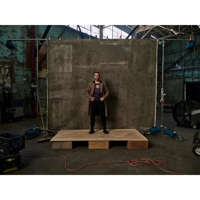 Full-length shot of an adult man standing on a wooden platform, wearing a jacket, t-shirt, and pants against a textured grey wall backdrop.