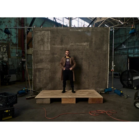 Full-length shot of an adult man standing on a wooden platform, wearing a jacket, t-shirt, and pants against a textured grey wall backdrop.