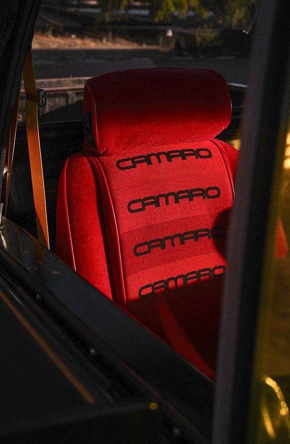Close-up of a red car seat with the word 'Camaro' stitched in black, seen through the vehicle's window with outdoor setting in the background.