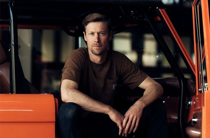 Adult male posing inside orange vehicle with dark interior. He is wearing a brown t-shirt and jeans while inside the car.