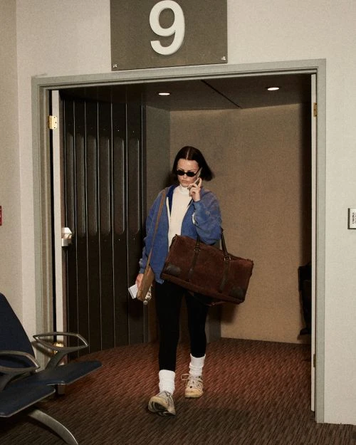 Adult female walks through a doorway in sunglasses, carrying a bag and talking on a cell phone, wearing an athleisure outfit.