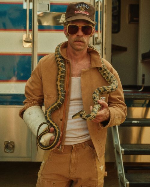 Man with snake draped around his neck stands outside with mobile trailer in background, wearing a jacket, cap, and sunglasses.