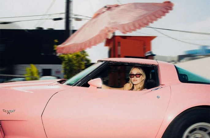 A fair-skinned adult female with sunglasses driving a classic pink Corvette convertible, with a blurry urban background.