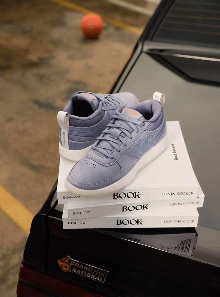 Nike sneakers displayed on Devin Booker books atop a black car, with a blurry basketball in the background, creating a sporty product shot.