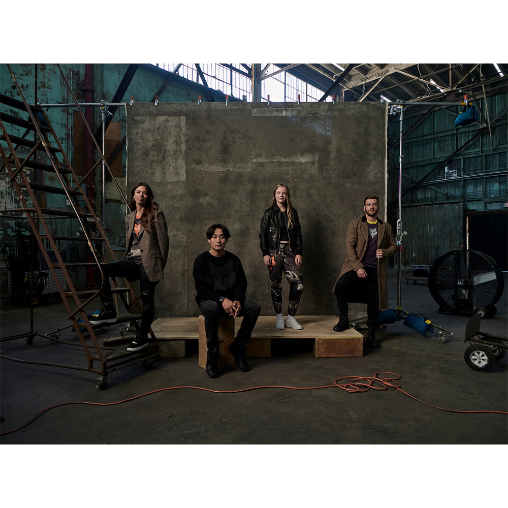 Group of four casually dressed adults in an industrial interior setting, posing on wooden platforms with film equipment nearby.