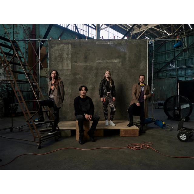 Group of four casually dressed adults in an industrial interior setting, posing on wooden platforms with film equipment nearby.