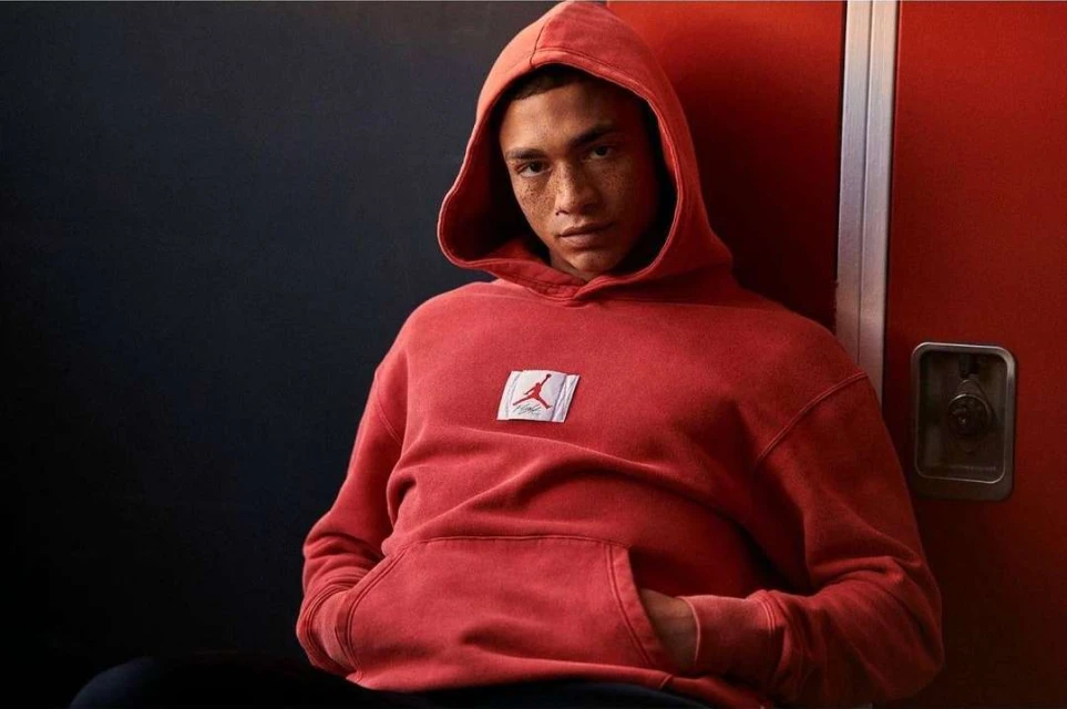 A young adult male wearing a red hooded sweatshirt with the Jordan logo poses against a red and blue wall indoors, looking at the camera.