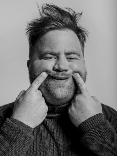 Close-up portrait of a Caucasian man with a beard and messy hair pulling the corners of his mouth up into a funny face against light gray.