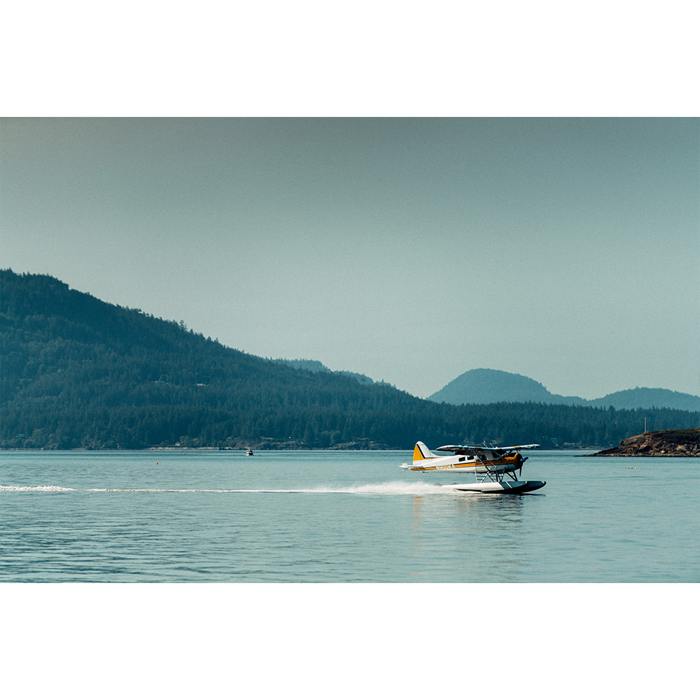 Seaplane takes off from a calm ocean surface, creating a trail of water spray, with forested mountains in the background.