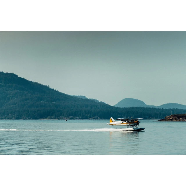 Seaplane takes off from a calm ocean surface, creating a trail of water spray, with forested mountains in the background.