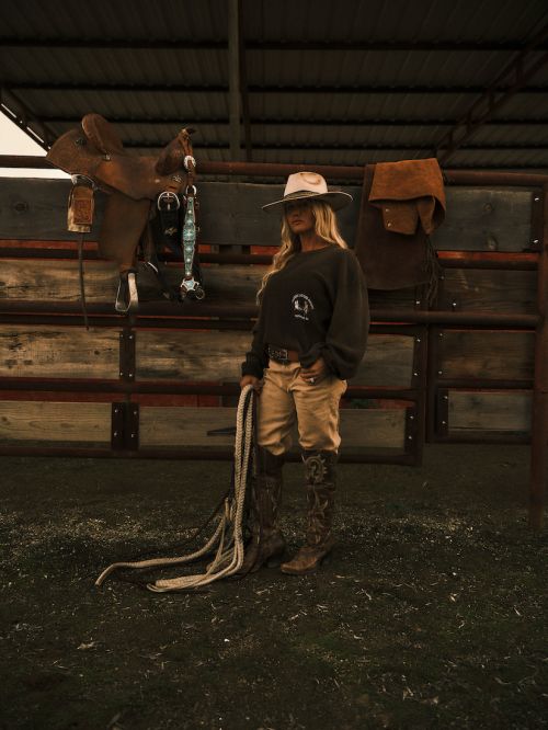 A woman wearing a cowboy hat and boots stands in a barn with a rope. A saddle and other tack hang on the wooden fence.