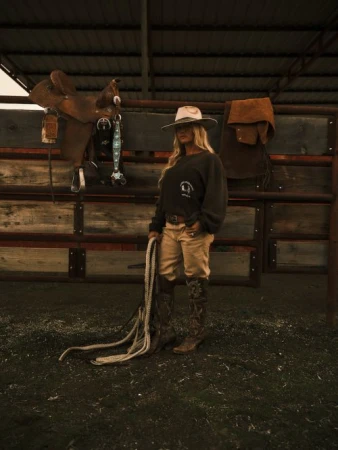 A woman wearing a cowboy hat and boots stands in a barn with a rope. A saddle and other tack hang on the wooden fence.