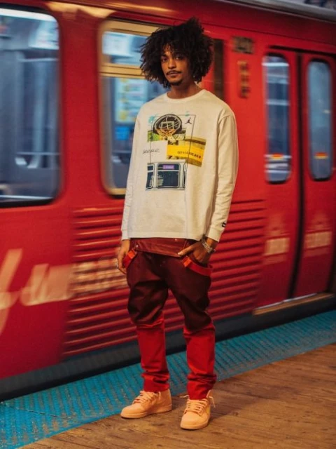 A slender man in a graphic tee and reddish-brown streetwear stands on a subway platform in front of a blurred red train.