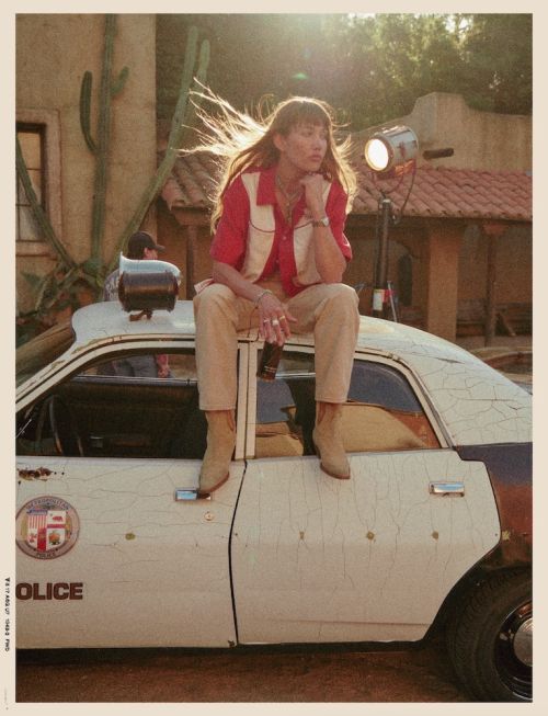 Woman with long hair sits on a weathered police car in a desert-like setting, illuminated by a light stand and backed by a building.